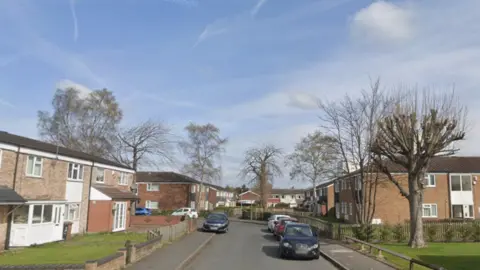 A residential street with brick houses, parked cars and a few trees under a blue sky.