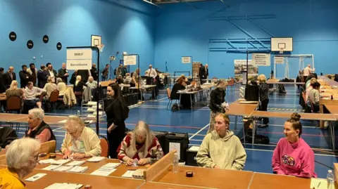 Women sit at desks in a sports hall counting ballot papers