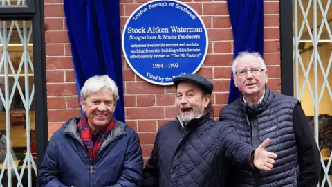 PA/Ian West From left to right, Mike Stock, Matt Aitken and Pete Waterman outside their former studios in London. Behind them is a blue plaque marking the site of headquarters, known as The Hit Factory.