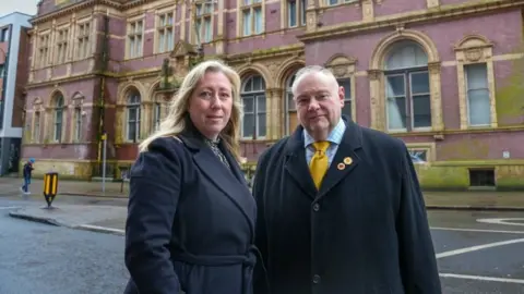 A woman with long blonde hair and a man with short greying hair are both wearing long black coats. They are standing in a city street in front of an ornate brick building with honey-coloured arched windows throughout.