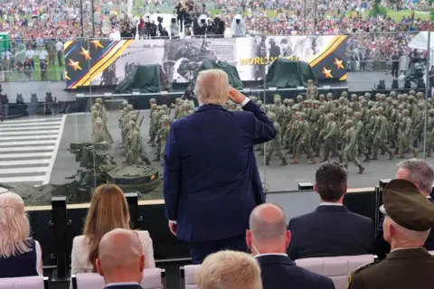 Reuters Trump is standing and facing the parade, his back turned to the camera and his right hand held to his right temple in a salute. Other dignitaries are sat around him and In front of him is the parade - dozens of soldiers marching past.