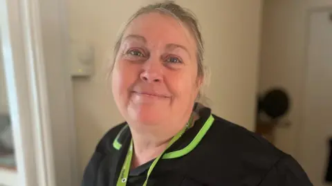 Kellie is smiling at the camera. She has blonde hair tied back. She is wearing a black work tunic that has like green trims.