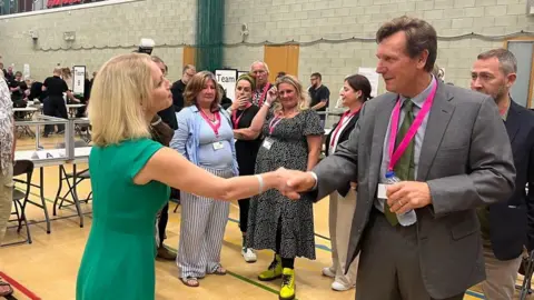 Charlotte Holloway, Labour councillor and co-leader of the Plymouth Knows Better campaign, in a green sleeveless dress, shaking hands with Angus Forbes from the Mayor for Plymouth campaign in a grey suit with a grey shirt and green tie and a pink lanyard and holding a water bottle.