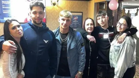 Cara Melia Wendy Chadwick is pictured with her two daughters and three sons. They appear to be in a pub. They are all smiling. One of Ms Chadwick's sons has his arms around his mum and one of his sisters. Another of her son's has his arm around one of her daughters.