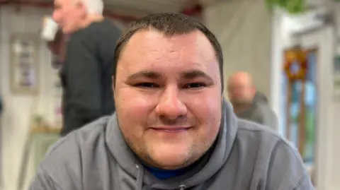 Jay Street is a 26-year-old man with short black hair. This portrait is head and shoulders. He wears a gray hoodie and is depicted in a cafe setting in rhyh, with people mingling behind him.