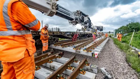 Six workers wearing orange high-vis uniforms and white helmets are working on laying a new piece of rail track onto the stones below. A metal crane is lowering the grey and copper tracks using red ropes. There is greenery bordering either side of the tracks.