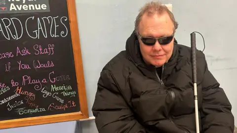 Dave Cunningham is wearing dark glasses and a black coat, and has a white cane in his hand. He is sitting sitting against a grey wall. There is a blackboard on one side and a bookcase on the other.