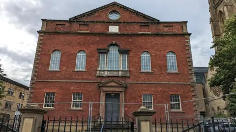 An 18th Century building in red brick over two storeys, with a a series of large windows in rows on two floors and a main doorway with stone steps leading to it