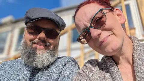 Sally Jones A couple smile as they look down into the camera. A man is on the left, a woman on the right. The man is wearing a blue cap, dark glasses and grey-and-blue patterned jumper. The woman has dark hair pulled back from her forehead. She is also wearing sunglasses. She has a green-and-white patterned cardigan. The picture has been taken on a sunny day. There is a building in the background, which is out of focus.
