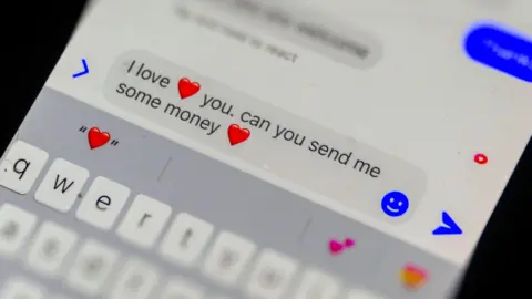 A mobile phone screen open to a messaging app with a incoming message on it which reads: "I love you. Can you send me some money." There are heart emojis on the message.