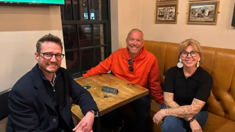 A man in bright orange hoodie sits at a reserved pub table with two others, a man and a woman, who are both wearing glasses