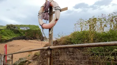 A lifebelt on a stick with some rope next to a metal fence leading to a beach beyond.