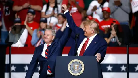 AP President Donald Trump and Senator Lindsey Graham are wearing navy blue business suits and standing behind a podium with the US presidential seal holding, each holding an arm up toward the other in a victorious gesture 