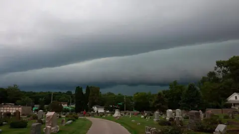  A person walks through Rose Hill Cemetery as a shelf cloud approaches behind them ahead of a severe thunderstorm. The clouds are grey and heavy in the sky. 