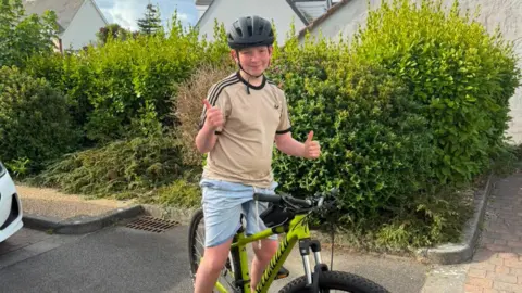 A boy on a push bike is smiling at the camera. He is wearing a safety helmet and is doing the thumbs up sign with both hands. He is in front of a large green hedge an appears to be in a car park near some houses. 