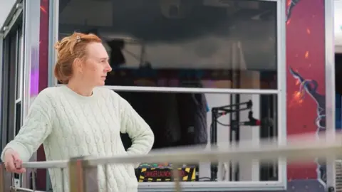 rachael wearing a white jumper and with red hair in front of a pay booth looks off camera to the right