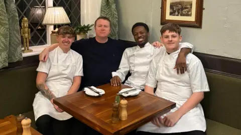 The three chefs are wearing their white cooking uniform and are sat with Mike on a corner table in the pub. Mike is wearing a dark jumper and has short brown hair. The seating is olive green with green curtains behind and a picture on the wall. They have a small square table in front of them.