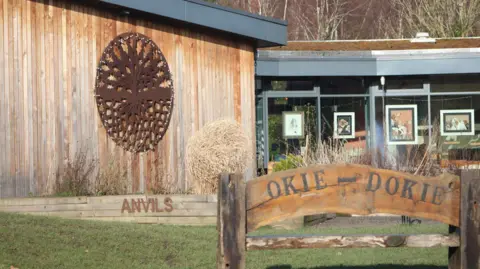 Groundworks NE & Cumbria A close up of the Land of Oak & Iron heritage centre. A wooden bench is outside and iron artwork of an oak tree hangs on an outer wall. Paintings of dogs are displayed inside the centre, which can be seen through it's windows.