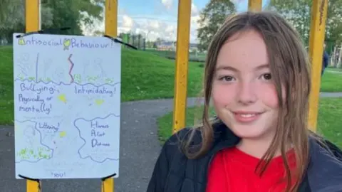 10-year-old Mia with brown hair, some of it down some of it tied up smiling into the camera, wearing a black coat and red jumper. She is standing next to her anti-social behaviour sign which is tied to bars in the park.