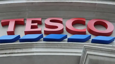 PA Media Tesco's logo as seen in front of a store. Large red fonts with blue dash outlines it.