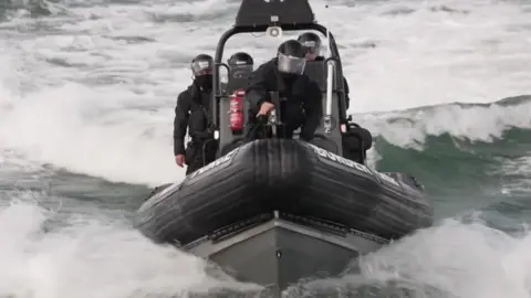 A black Rib sails towards the camera creating a bow wave in the water. There are four people onboard all wearing black with helmets and visors.