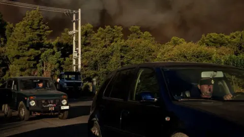 People drive as smoke rises from a wildfire in the village of Barnabas, near Athens, Greece, August 11, 2024. REUTERS