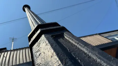 Jodie Halford/BBC A tall, black, cast iron pipe stretches up from the bottom of the photo towards a blue sky. The pipe has indentations and decorative features which made it look Victorian. A house and some TV aerials and cables are at the top of the picture.