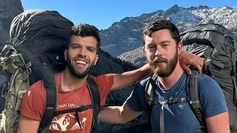 Aziz Ziriat, left, and Samuel Harris in shorts, t-shirt wirth large backpacks in front of a lake surrounded by mountains