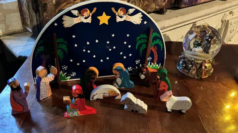 A small wooden nativity scene on a wooden table top. Painted wood in a simple style, a background with two angels around a star on a night sky. The figures in front are separate and movable, all in traditional dress but cartoon-like. To the right is a snow globe with a separate nativity inside.