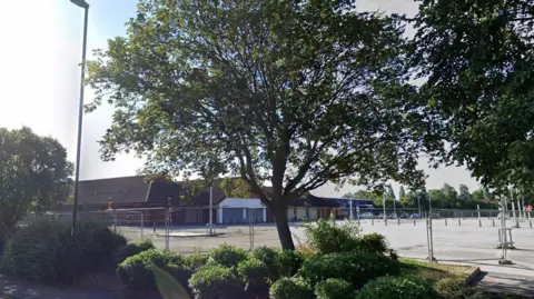 A boarded up supermarket building with barriers around the car park. The site stands empty. There are trees and shrubs planted around the site.