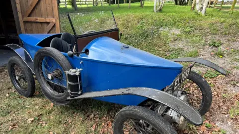 Humbert & Ellis An Alcyon Cyclecar resting on grass strewn with brown leaves. The vehicle has a bright blue body, no roof and black tyres and hub caps. Behind its windscreen are two seats. 