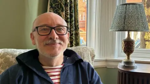 Andy Hughes sitting in front of his front window at home in Nuneaton. A lamp sits on the table behind him and neighbouring homes and parked cars can be seen outside the window in the morning sunshine.