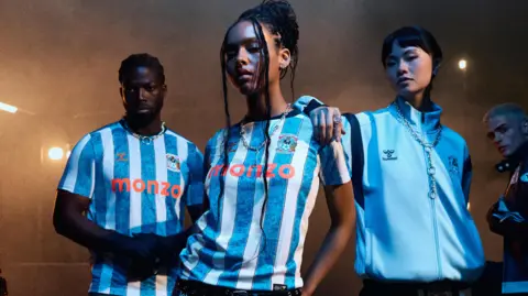 Coventry City Football Club Male and female models are wearing the new Coventry City kit. It is blue and white striped, with a pattern in the blue stripes, the club crest on the left breast and the sponsors name across the middle.