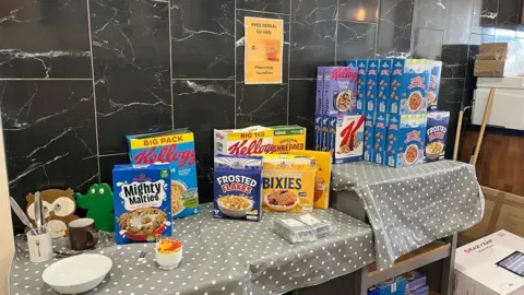 Lucinda Adam / BBC A table with a number of boxes of cereal on, as well as a grey polka dot tablecloth. A sign on the wall reads "Free cereal for kids. Please help yourselves".