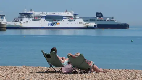 PA Media Two women reclining on beach chairs on a sunny pebble beach. Two large ferries can be seen at sea near the shore.