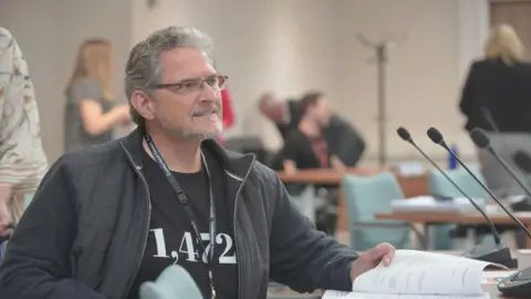 Brian Wernham with grey hair and beard sitting down with a black jacket and T-shirt. He is flicking through a document. 