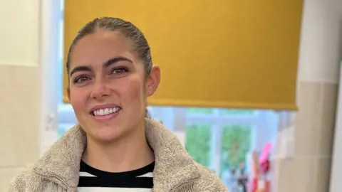 Molly smiles at the camera as she stands by a kitchen counter with two cake mixers to her left. She's wearing a white fleece with a black and white stripped t-shirt. Her hair is tied back in a ponytail.