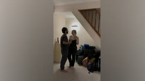 Billie Van Der Walt A man and woman, dressed in dark clothing, hold two wine glasses as they cheers each other. Both are standing in an empty living room, lit by a wall light and surrounded by bags and backpacks.