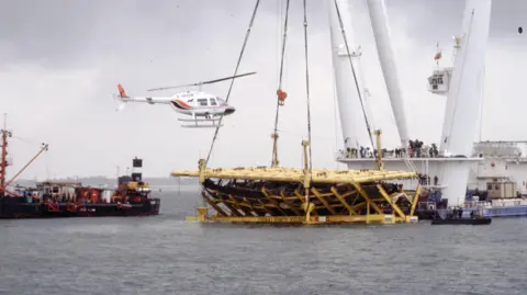 Getty Images A ship is pictured hoisting something large out of the water with a big crane. Nearby is another ship and a helicopter hovering low in the sky. 