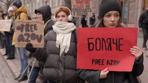 People can be seen at the protest, including a lady holding a red sign "Free Azov"