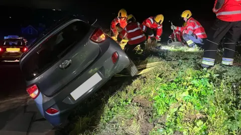 Jersey Fire & Rescue Service A grey car mounted on a mound of grass at a 45 degree angle, with fire officers standing on the mound holding onto the car by anchoring it with equipment.