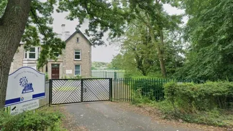 The gates of the Meadows School. It is a large three-storey grey brick house with a brown door. It is surrounded by trees and shrubs.