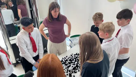 BBC Seven young secondary school pupils and a teacher standing in front of desk which has some black and white balls on it which represent the HPV virus