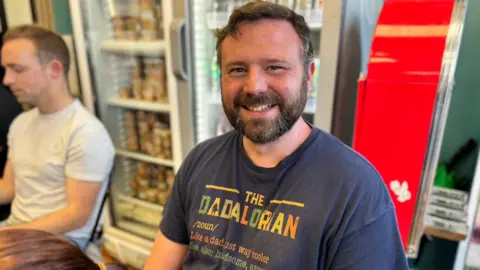 Karl Boyle has short dark hair and a short beard. He's sitting in a cafe in front of a drinks fridge and is wearing a navy blue t-shirt that says "the dadalorian" in colourful lettering. He is smiling. 
