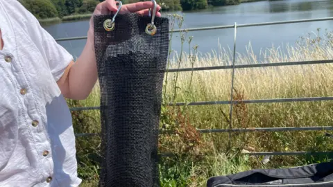 A mesh bag containing black pellets being held up to the camera. A reservoir is visible in the background. 