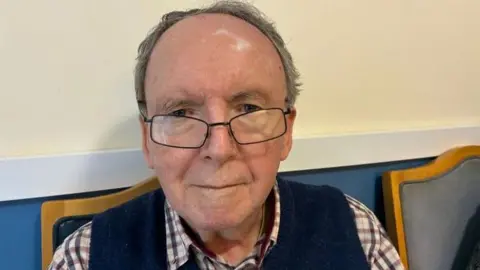 Richard is an elderly gentleman smiling slightly at the camera. He has lost most of his hair and wears thick spectacles with a navy tank top over a white, burgundy and navy chequered shirt. He is sitting on a wooden chair with a blue leather backrest, up against a cream wall. 