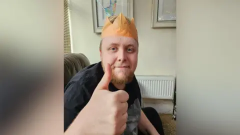 Samantha Fairbairn Andre Yarham wears an orange hat from a Christmas cracker and is looking at the camera and giving a thumbs up with his hand.