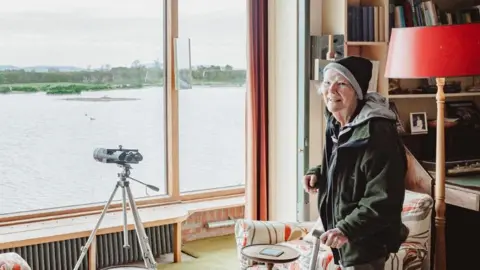 An elderly woman in a hat stands by a large window with a telescope in front of it