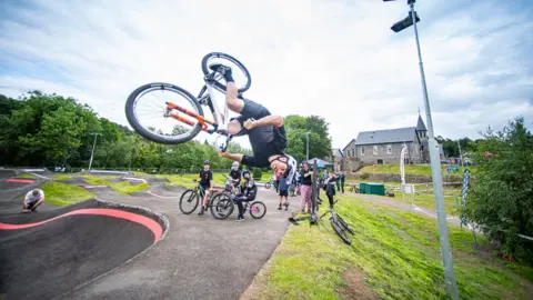 A bike rider is upside down over a track with other riders looking on and a large granite building in the background.