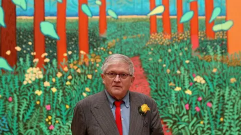 A man with grey hair and wearing black-rimmed glasses is standing in front of a colourful painting depicting green plants and tall trees. He is wearing a grey pin-striped suit, blue shirt and red tie with a yellow flower in his lapel. 